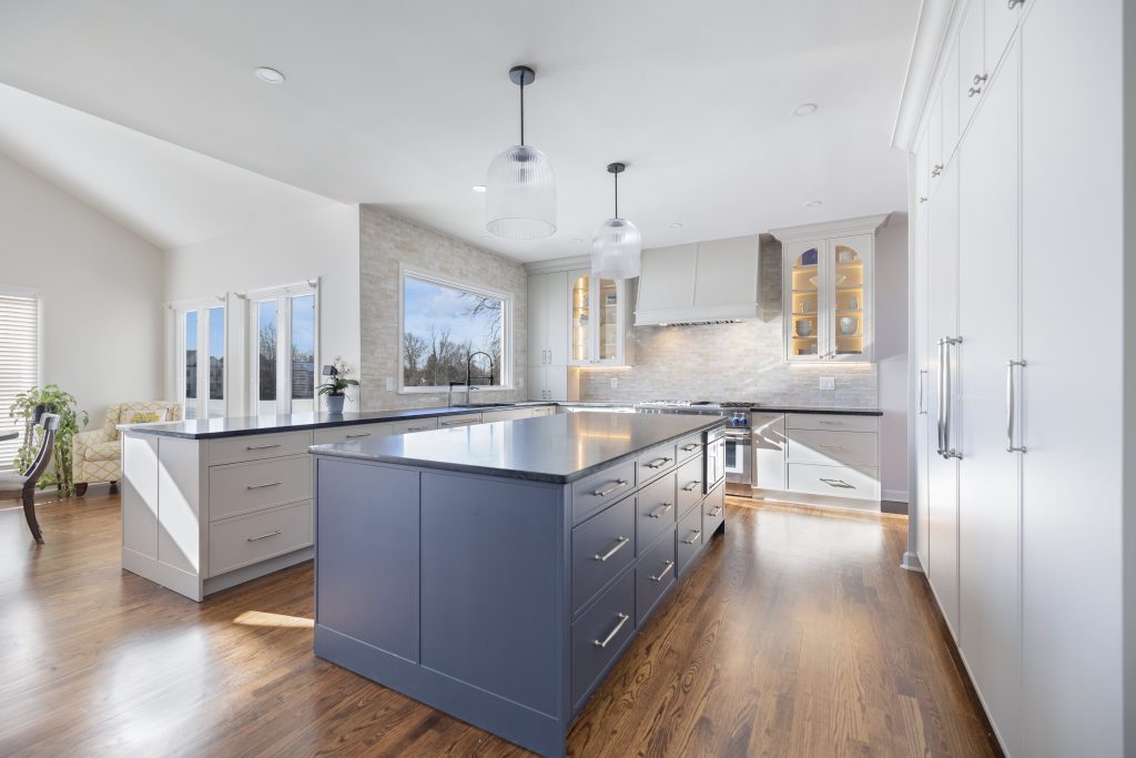 Modern kitchen with white cabinets, a large navy blue island, pendant lights, stainless steel appliances, wooden floors, and large windows letting in natural light.