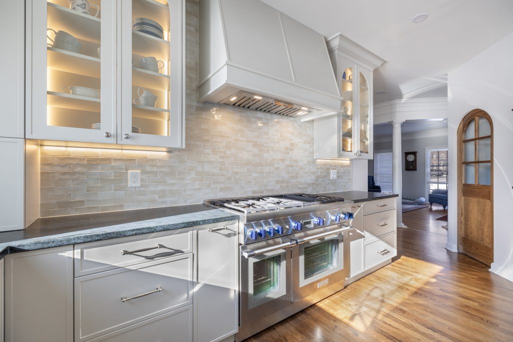 Modern kitchen with white cabinets, open shelving displaying dishes, a stainless steel gas stove with blue knobs, stone tile backsplash, under-cabinet lighting, and wood flooring illuminated by natural light.