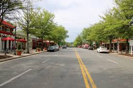 A quiet, empty street lined with trees and small shops on both sides. A few parked cars are visible, and red umbrellas can be seen outside some storefronts under a cloudy sky.