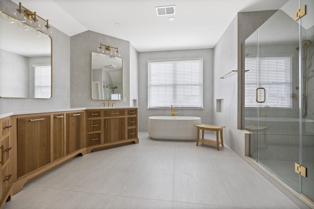 Spacious modern bathroom with light gray walls, a large wooden vanity with double sinks, two mirrors, a freestanding bathtub, a glass shower, a wooden stool, and a window with white blinds letting in natural light.