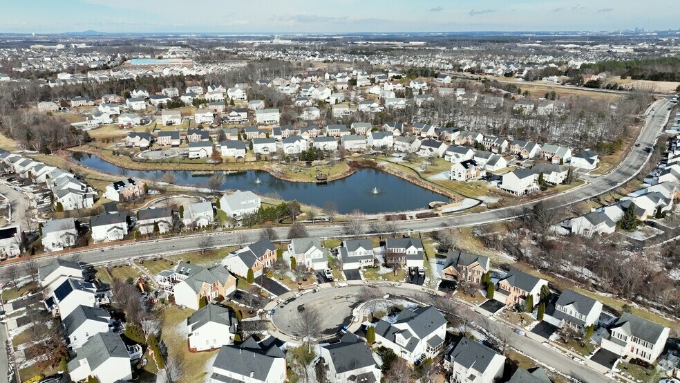 Aerial view of a suburban neighborhood with rows of houses, curved streets, a small pond in the center, and patches of trees and grass throughout the community.