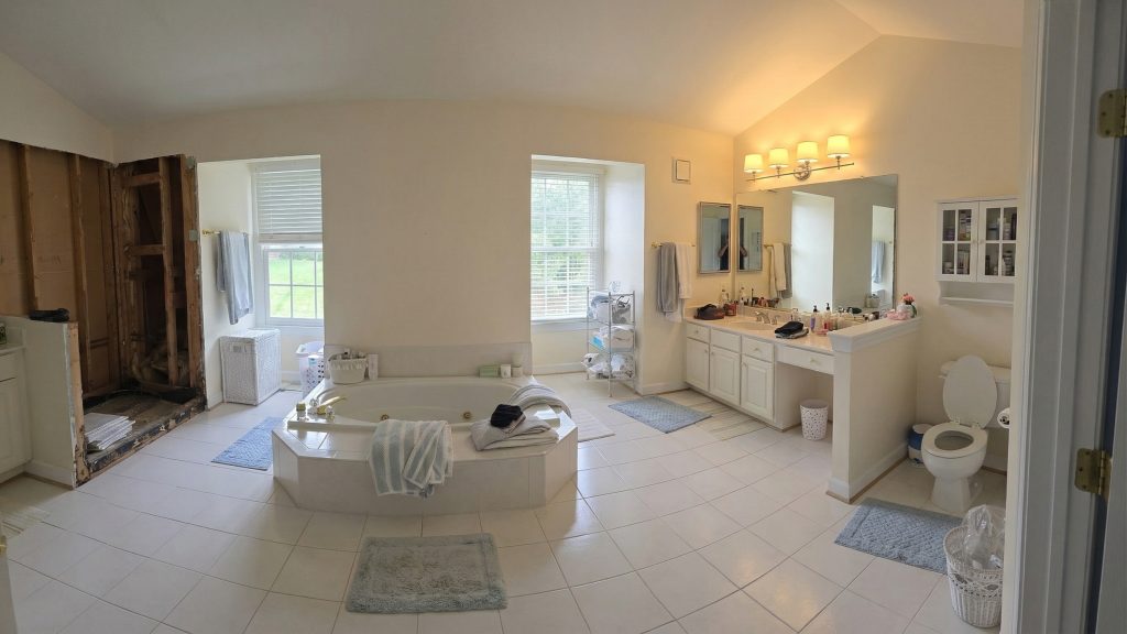 Spacious bathroom with white tile, a central bathtub, double sinks, toilet, large windows, mirrors, and blue rugs. The left side shows exposed wall studs and pipes, indicating partial renovation.