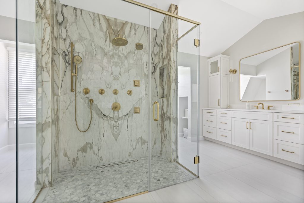 A luxurious bathroom with a large glass-enclosed shower featuring marble walls and gold fixtures, next to a white double vanity with gold hardware and a large mirror. Natural light streams in from a nearby window.