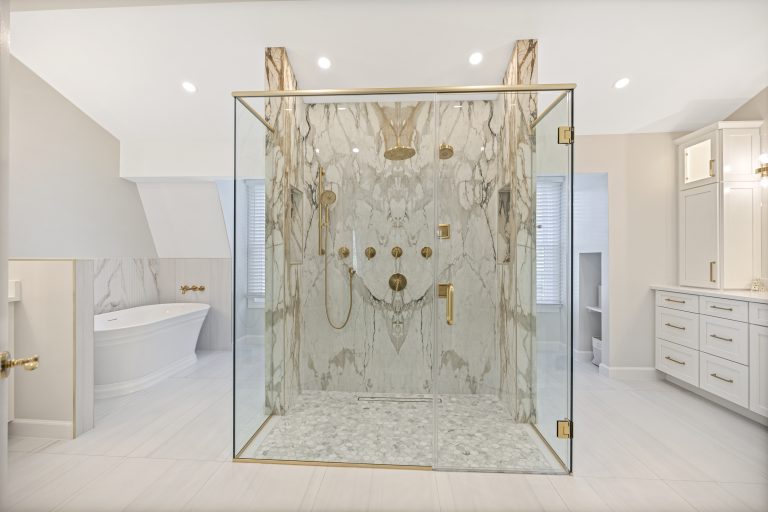 Luxurious bathroom with a glass-enclosed marble shower featuring gold fixtures, a freestanding bathtub with gold taps to the left, and white cabinets with gold handles on the right. Neutral tones create an elegant look.