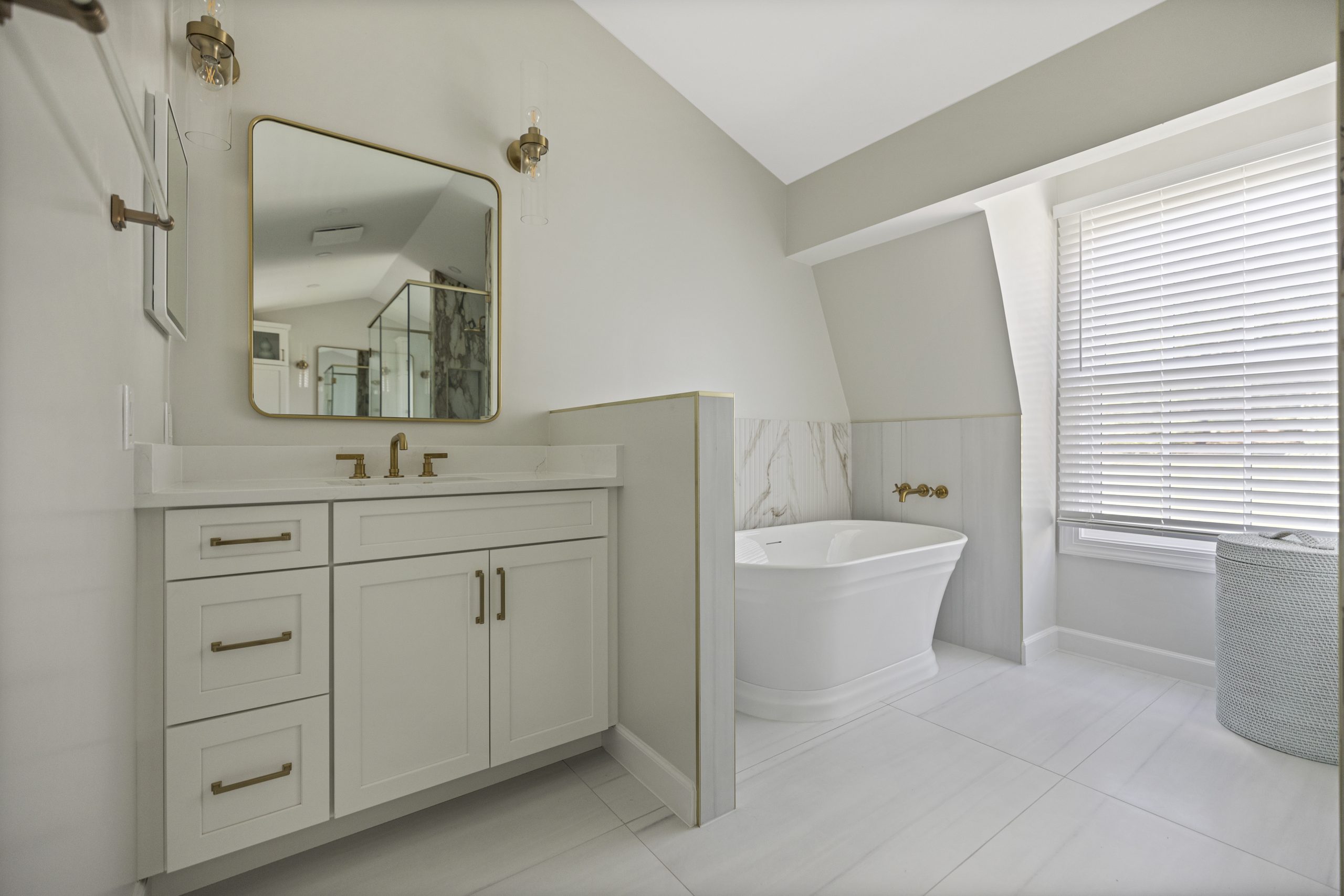 Modern bathroom with white cabinets, gold fixtures, a square mirror, freestanding bathtub, marble accent, large window with blinds, and a laundry basket on white tile floor.