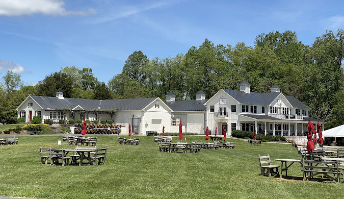 A spacious grassy lawn with several wooden picnic tables and red umbrellas sits in front of a large, white farmhouse-style building surrounded by green trees under a partly cloudy sky.