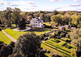 Aerial view of a large white mansion surrounded by green lawns, trees, and landscaped gardens under a partly cloudy sky. A circular driveway and formal hedges are visible in the foreground.