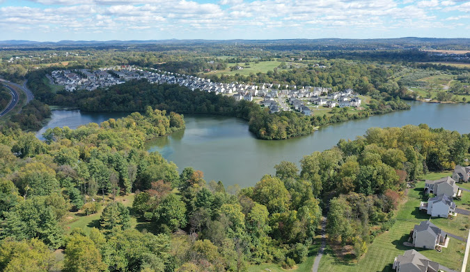 Aerial view of a residential neighborhood surrounded by trees and a large lake, with houses clustered near the water and stretches of green landscape in the background under a partly cloudy sky.