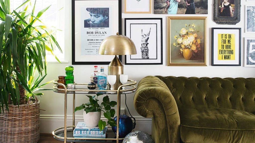 A green velvet sofa sits beneath framed art, next to a gold lamp, bar cart with drinks, and lush plants.