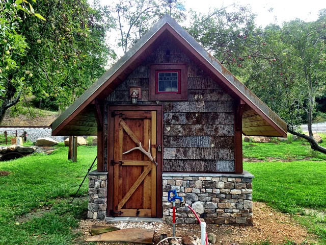 A small stone and wood shed with a shingled roof stands among grass and trees, wooden door visible.