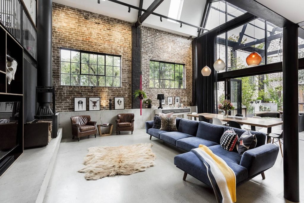 Modern loft with large windows, exposed brick, a blue sofa, leather chairs, and a dining area.
