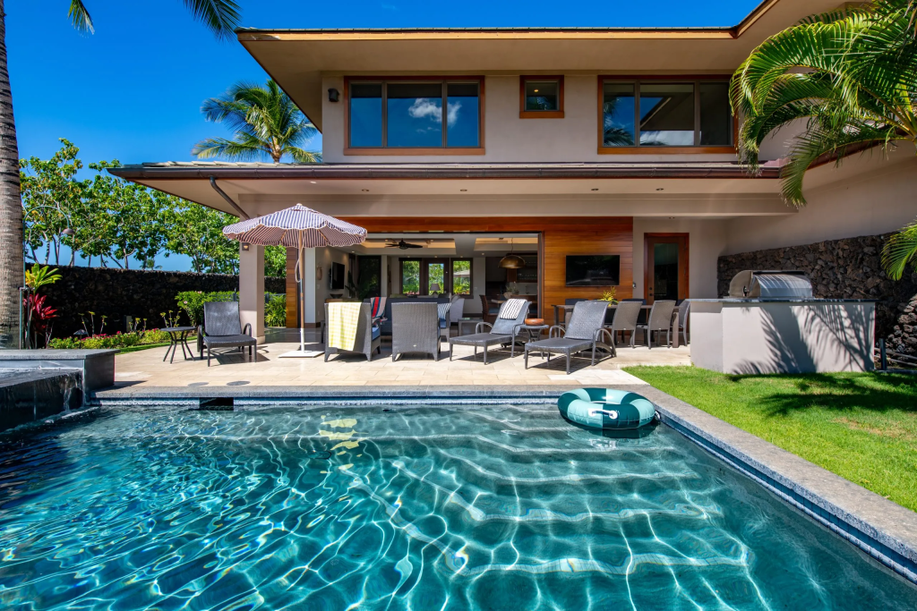 Modern house with a pool, patio chairs, and palm trees under a clear blue sky on a sunny day.