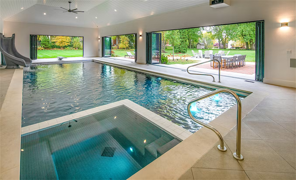 An indoor pool with a slide, hot tub, and open glass doors overlooks a patio with outdoor seating and greenery.