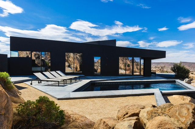 A modern black house with large windows and a pool sits in a rocky desert landscape beneath a blue sky.