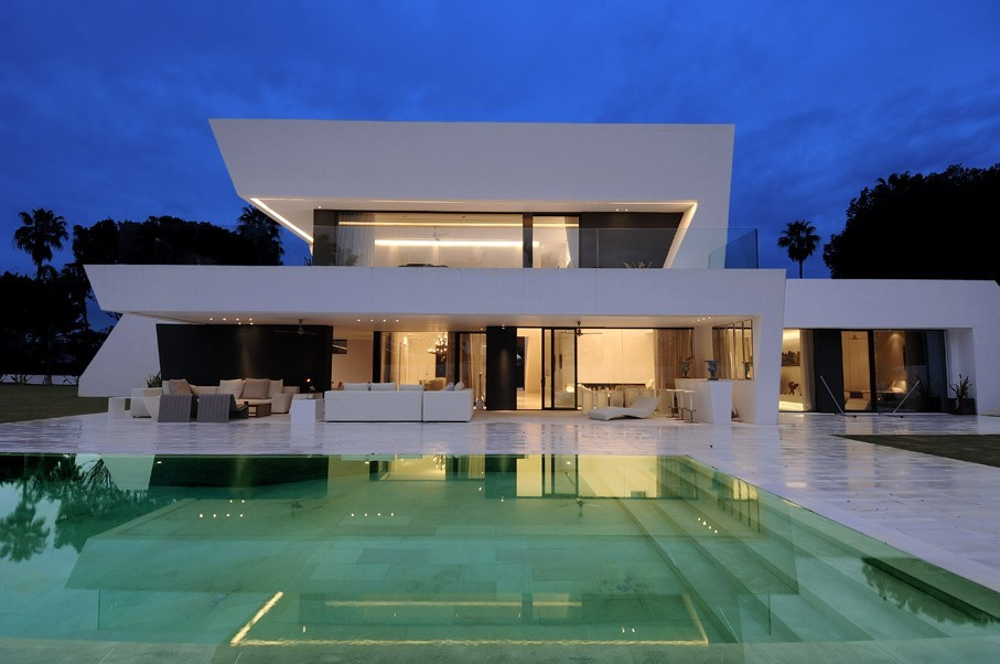 Modern white two-story house with large windows and a glowing swimming pool at dusk.