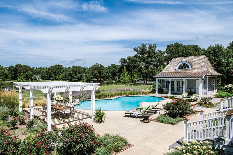 A backyard features a pool, lounge chairs, a pergola, flowers, and a small cottage below a blue sky.