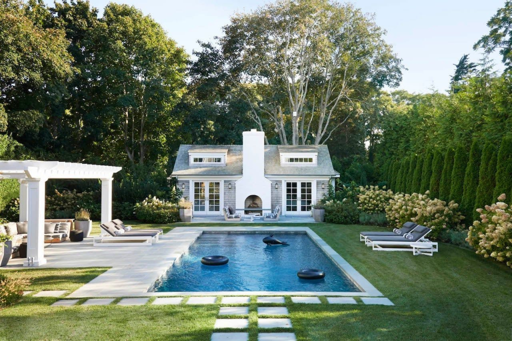 A backyard pool features floating tubes, lounge chairs, a white pool house, and lush green landscaping.