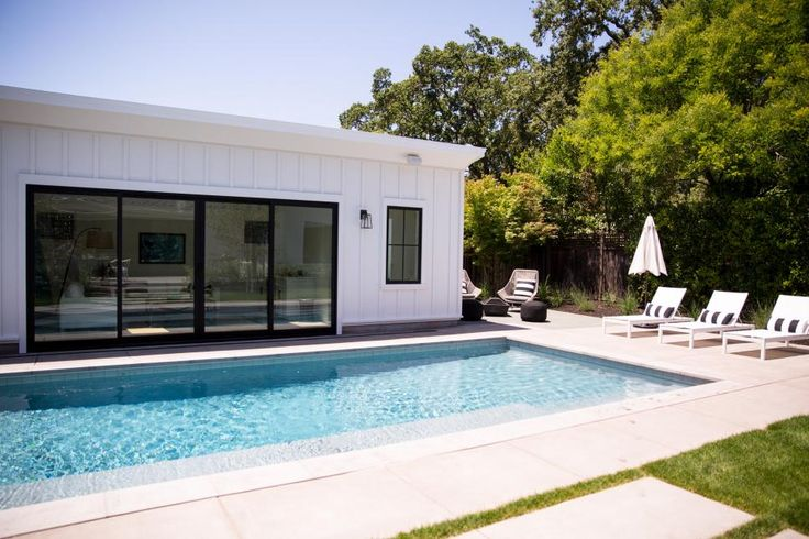 Modern backyard features a rectangular pool, lounge chairs, umbrella, and a white house with glass doors.