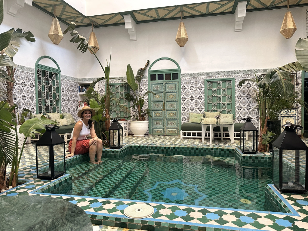 A woman in a hat sits by a tiled indoor pool surrounded by lush plants and Moroccan-style decor.