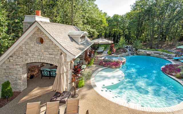 A backyard pool features a stone pool house, patio chairs, umbrellas, and lush landscaping by tall trees.
