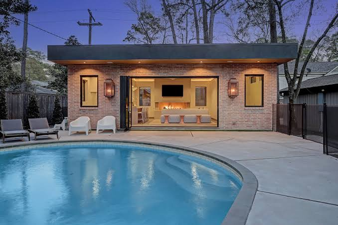 Modern pool house with brick exterior, open patio doors, lit fireplace, and a curved pool at dusk.