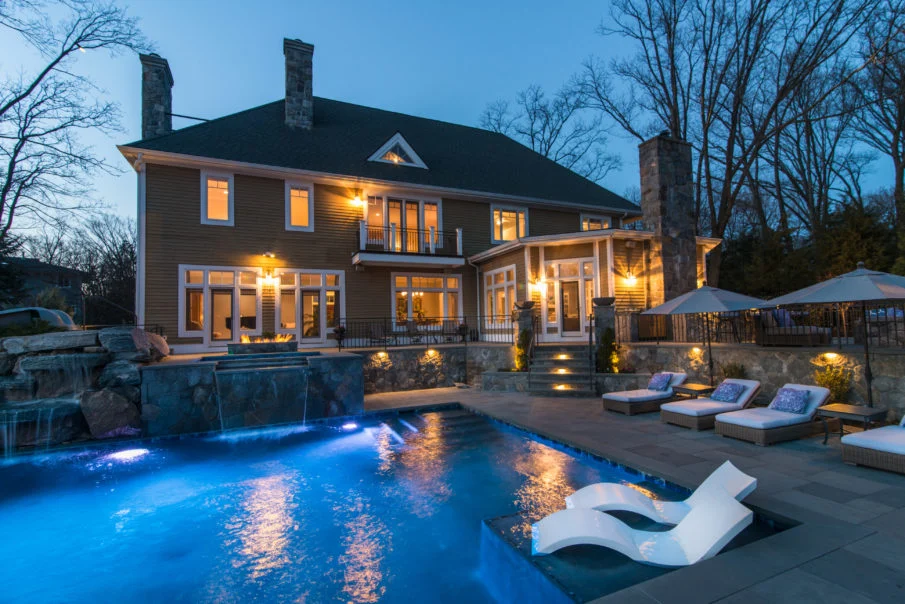 A large house with lit windows, a glowing pool, lounge chairs, and trees creates a serene dusk retreat.