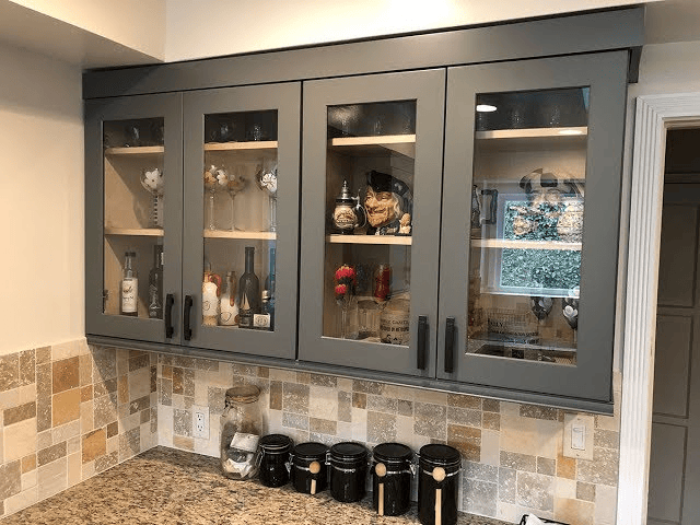 Gray kitchen cabinets with glass doors showcase glasses; black jars rest on a granite countertop below.