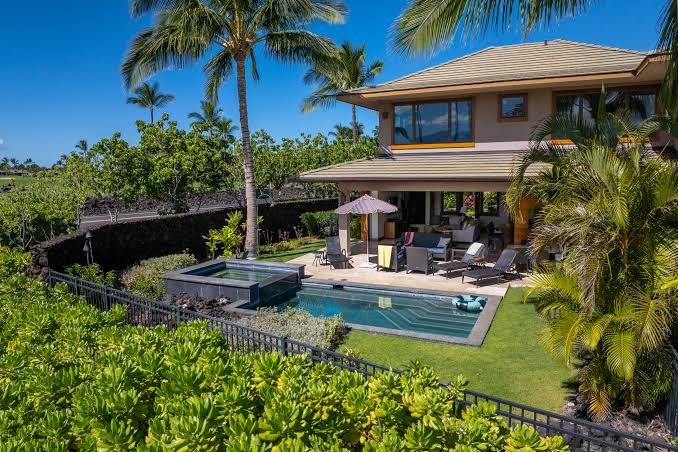 A tropical house with a patio, pool, hot tub, palm trees, and a lush green garden under a blue sky.