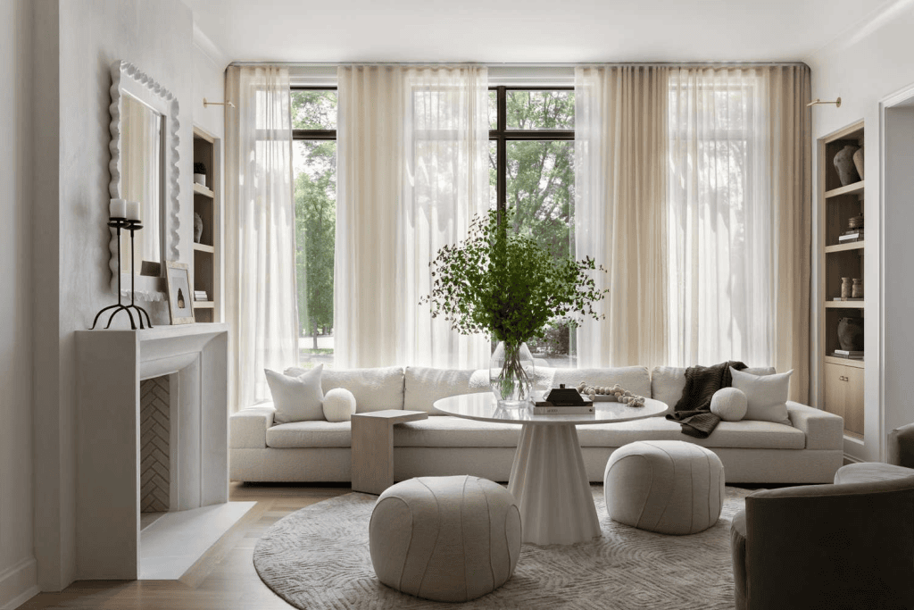 Modern living room with large windows, sheer curtains, neutral furniture, and a round coffee table with plants.