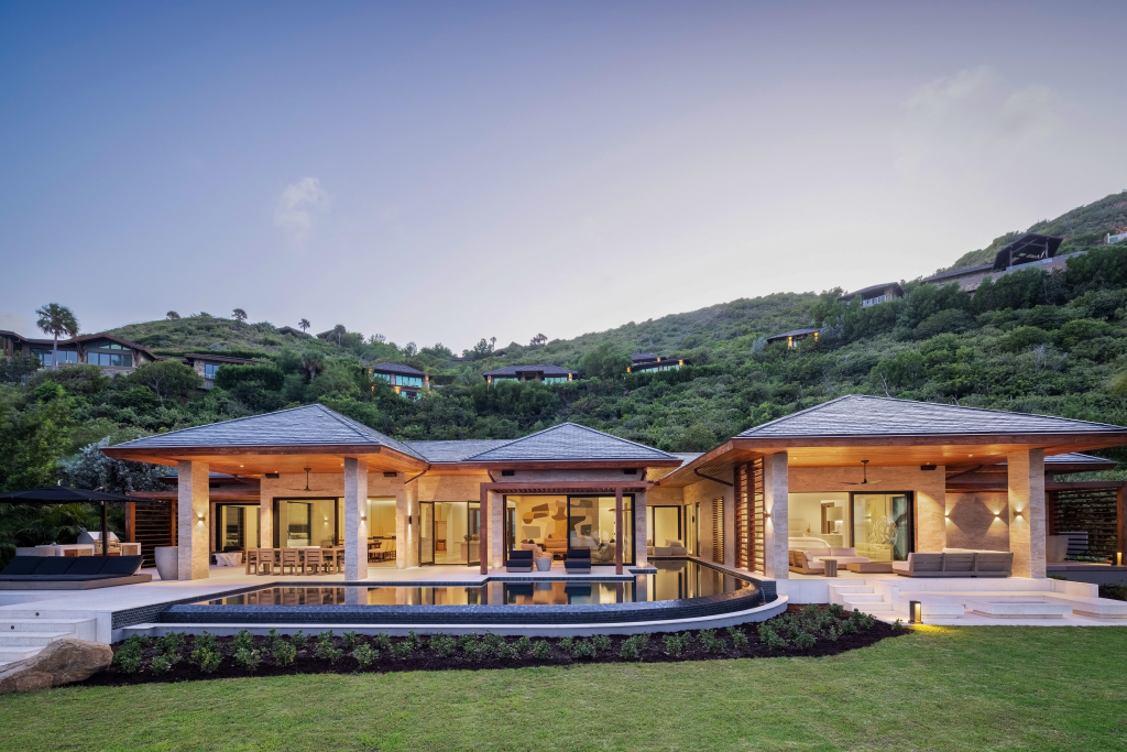 Modern luxury villa with large windows, pool, and patio overlooks green hills at sunset.