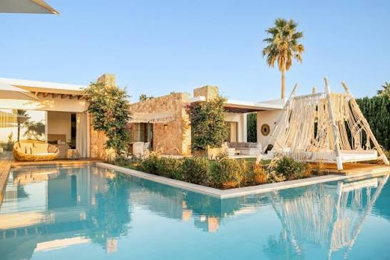 A modern villa with stone walls and a pool features a palm tree and large white outdoor hammock beneath a blue sky.