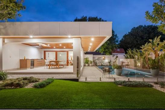 Modern backyard at dusk with outdoor kitchen, dining area, and pool surrounded by lush plants.