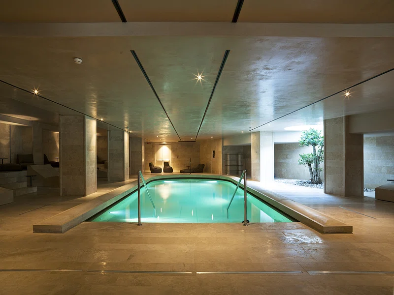 Modern indoor swimming pool with steps, beige stone walls and floor, softly lit for a serene atmosphere.