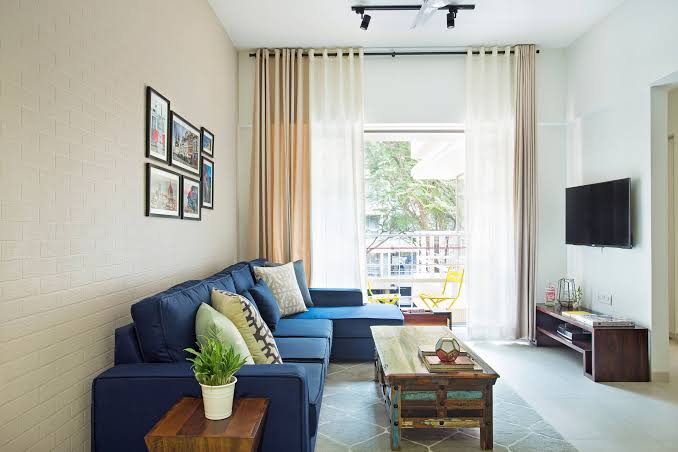 Bright living room with a blue sofa, coffee table, TV, wall art, plant, and large window with curtains.