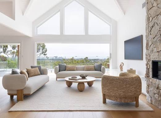 Modern living room with neutral sofas, round table, large windows, and a stone fireplace.
