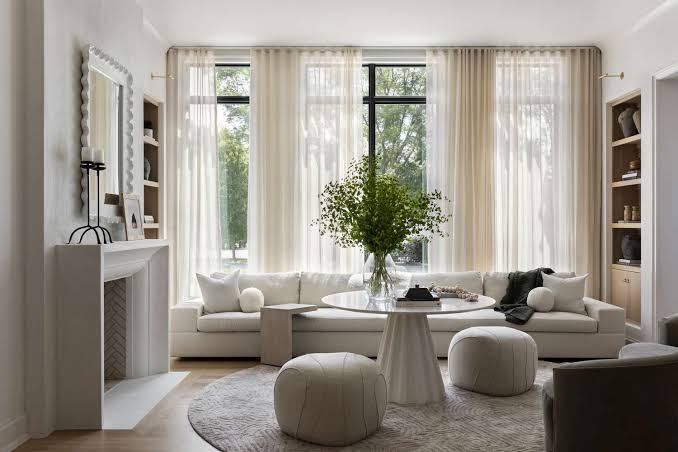 Modern living room with white furniture, round table, leafy centerpiece, large windows, and sheer curtains.