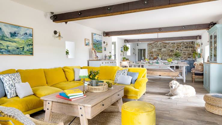 Bright living room with a yellow sofa, wooden table, books, and a dog lying on the floor.
