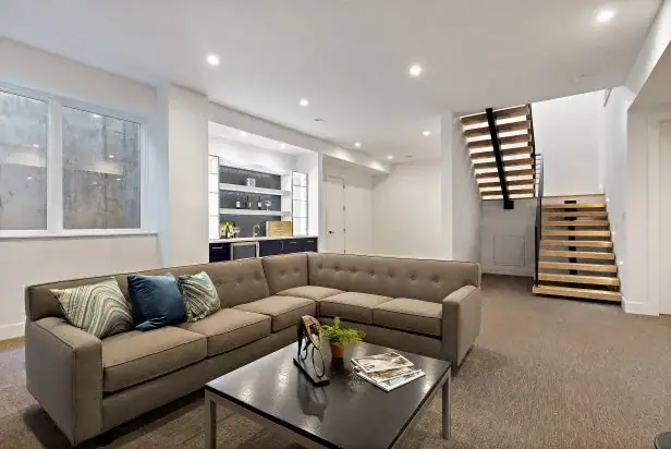 Modern basement features a gray sectional sofa, sleek coffee table, open staircase, and built-in bar area.