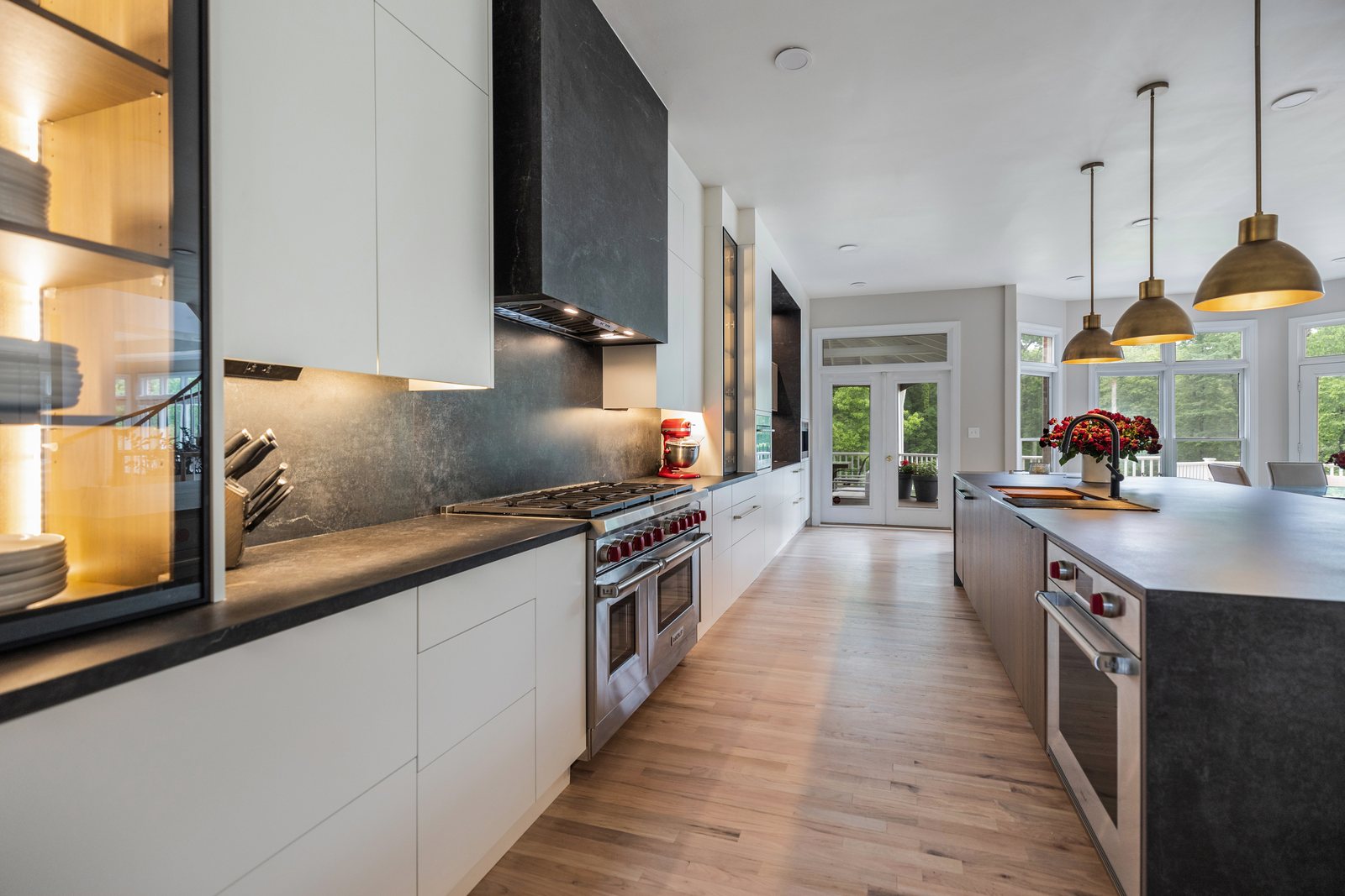 Galley style kitchen by EA Home Design in Leesburg: white flat panel cabinets with lit glass uppers on one wall, a black stone hood and backsplash over a professional range, and a long dark stone island with gold pendants as the second run