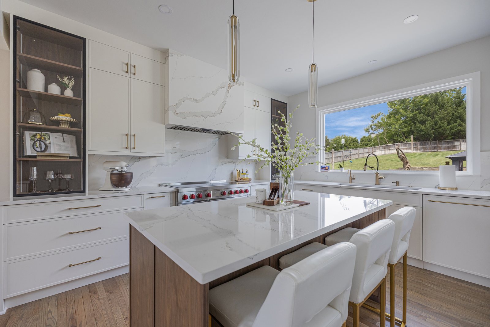 Open galley kitchen in Northern Virginia by EA Home Design with a white cabinet run, a glass-front upper, a professional range under a plaster hood, and a marble waterfall island with brass hardware and four stools