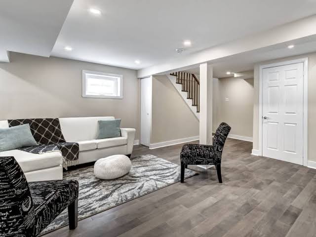 Modern basement with neutral tones features a white sofa, chairs, rug, and stairs leading upstairs.