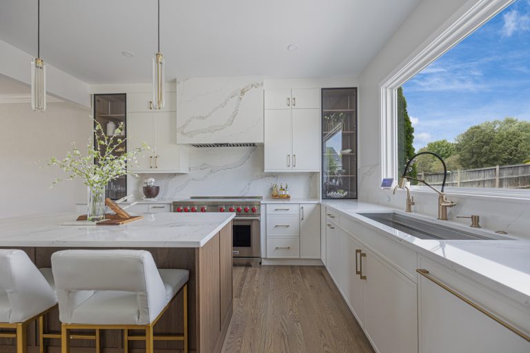 A white modern kitchen features a marble waterfall island, gold hardware, and a large window.
